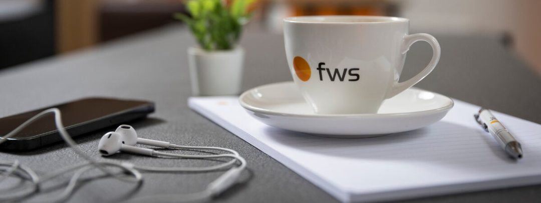 fws logós kávéscsésze jegyzettömbön, mellette tollal, telefonnal és fülhallgatóval