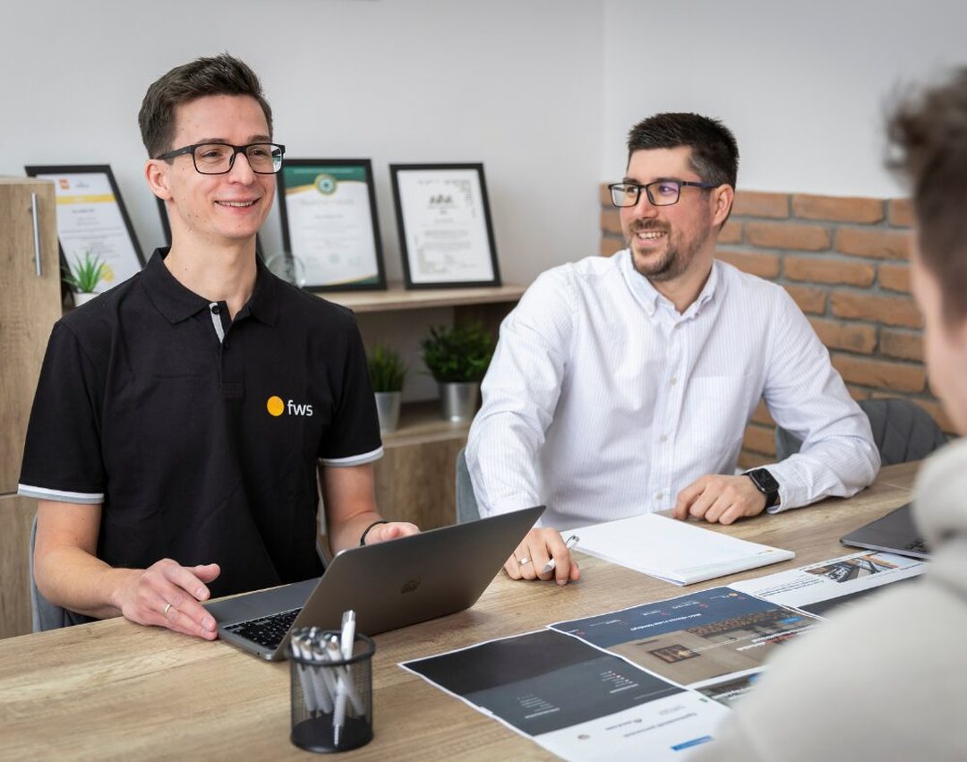 Az fws két szakértője ügyféllel egyeztet, az asztalon laptop és kinyomtatott weboldaltervek