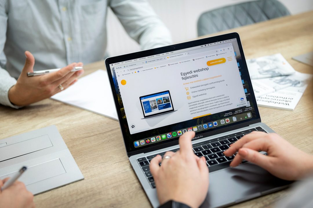 Laptopon az fws.hu „Egyedi webshop fejlesztés” oldala, mellette tollal a kezében magyarázó kolléga