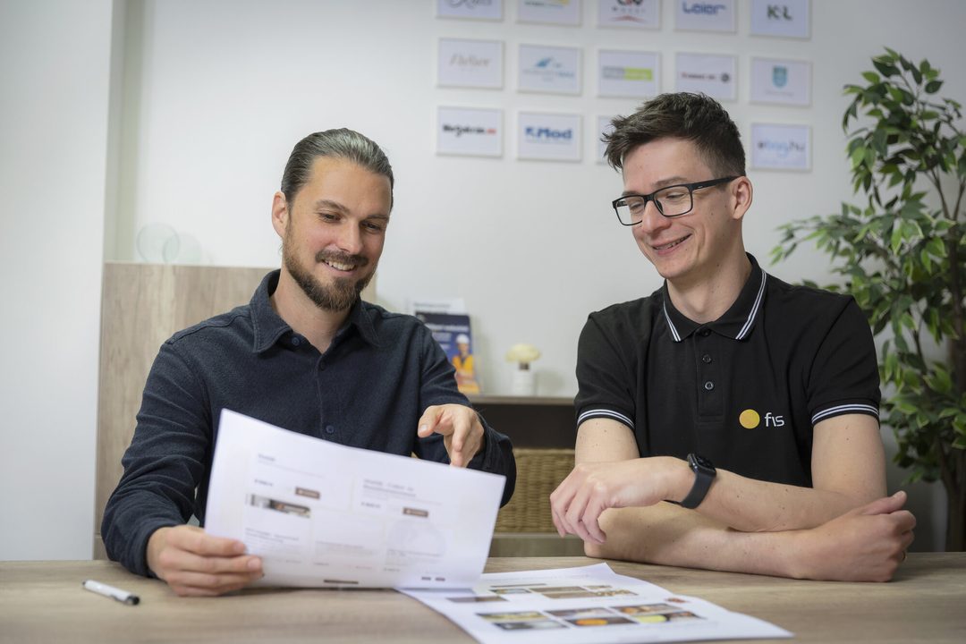 Két fws munkatárs kinyomtatott weboldalterveket néz át, a háttérben ügyféllogók a falon